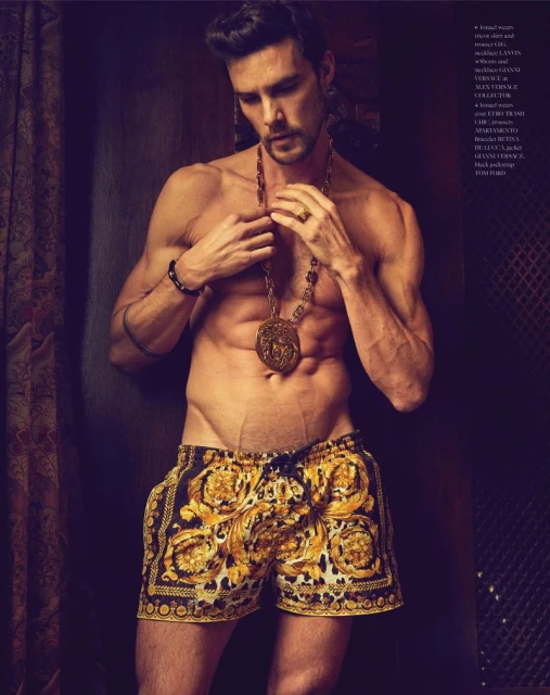 Male fashion model poses shirtless in Versace print shorts with gold jewelry and earrings against a dark studio backdrop.