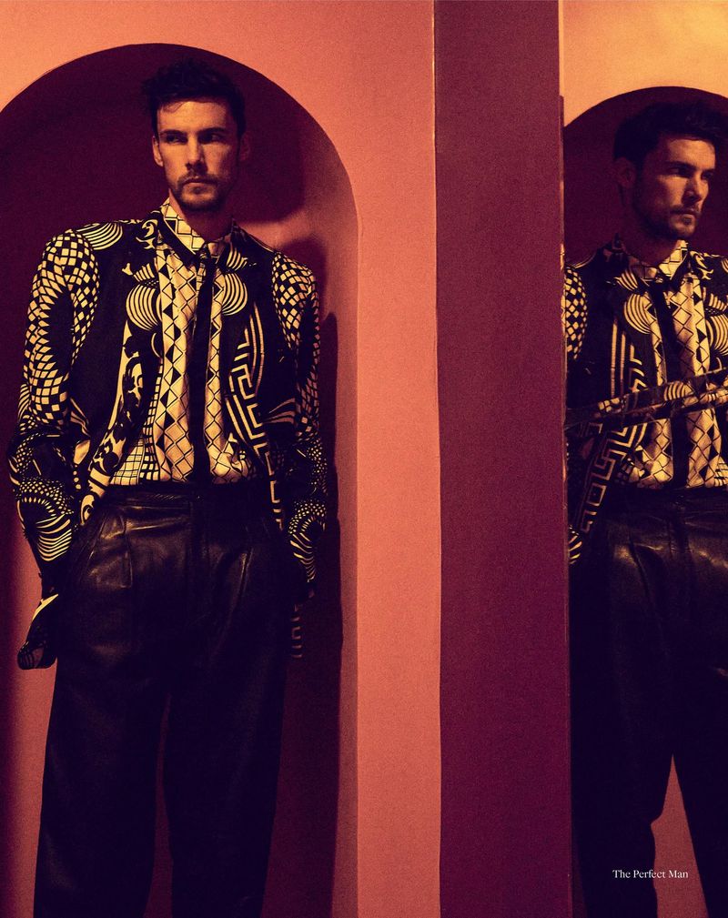 Portrait of a dark-haired male fashion model with a patterned jacket and leather pants, posed in a salmon-colored arched alcove.