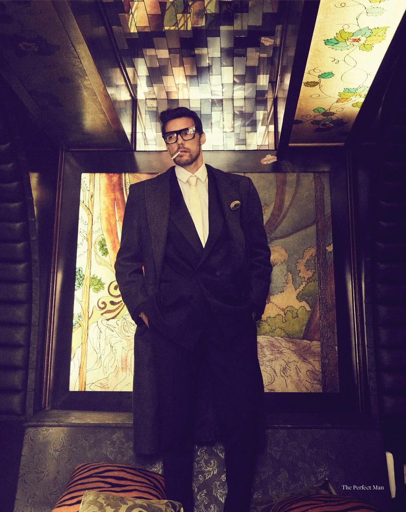 Mature man in suit and glasses smoking, posing in dark interior setting with ornate painting and patterned pillows.