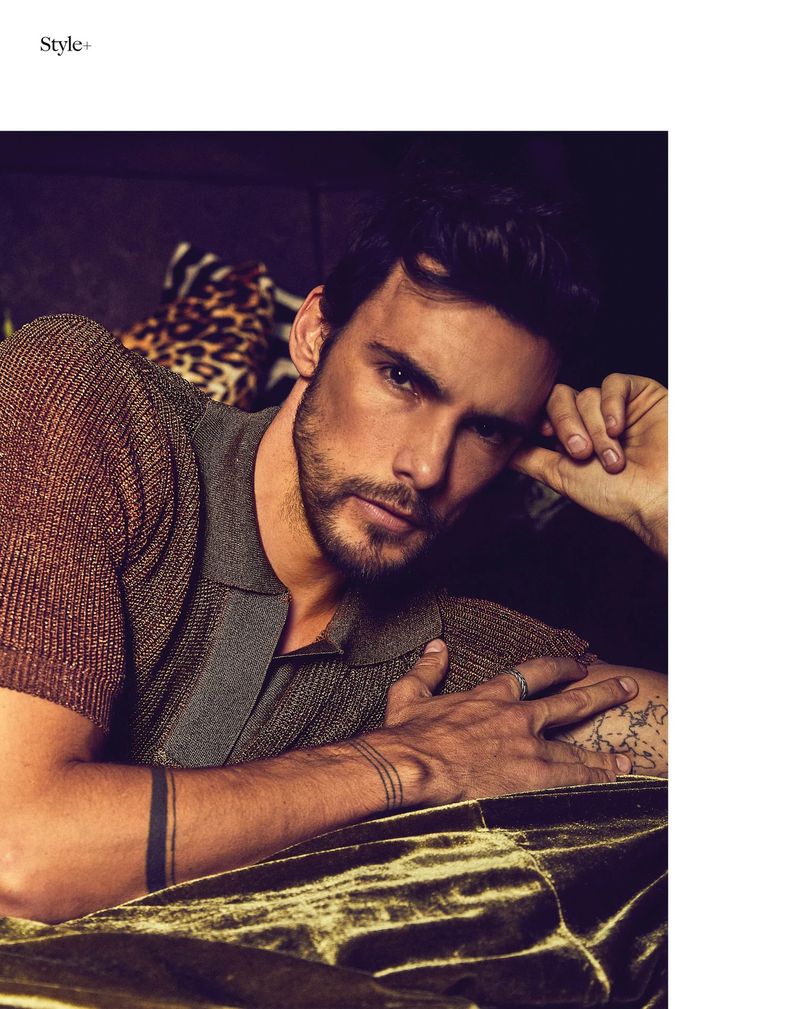 Portrait of a handsome adult male model with dark hair and a beard, laying on an olive velvet surface, wearing a bronze short-sleeve shirt.