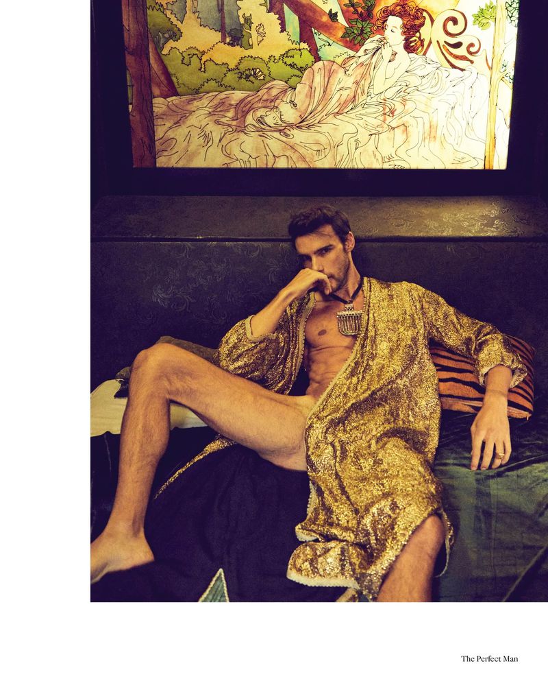 Attractive man lounges on a couch in a gold robe in a fashion editorial, featuring an artwork backdrop in an indoor setting.