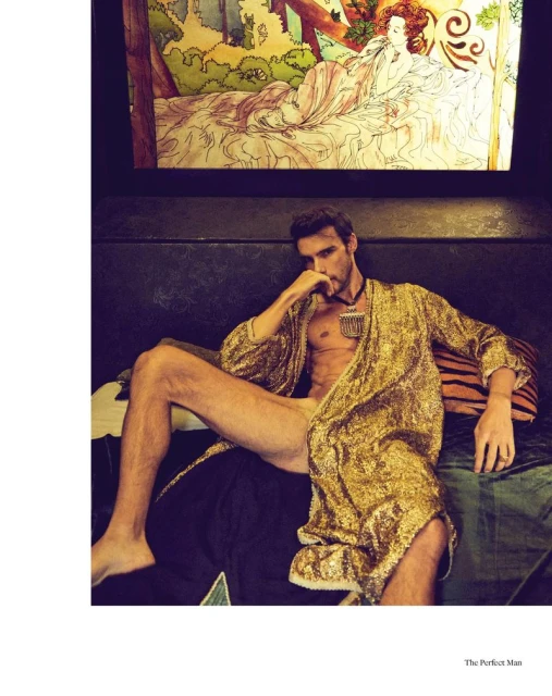 Attractive man lounges on a couch in a gold robe in a fashion editorial, featuring an artwork backdrop in an indoor setting.