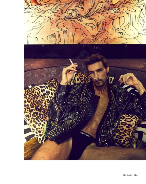 Adult male lounging on pillows in a black jacket smoking a cigarette. Man wearing branded underwear, black nail polish, and lying on a bed.
