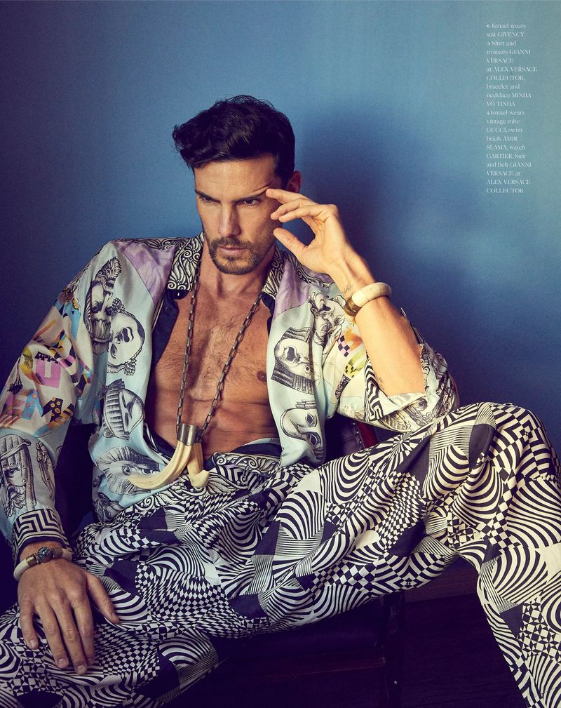 A stylish male model poses indoors wearing a patterned shirt and pants with ivory bracelets and a necklace, set against a blue background.