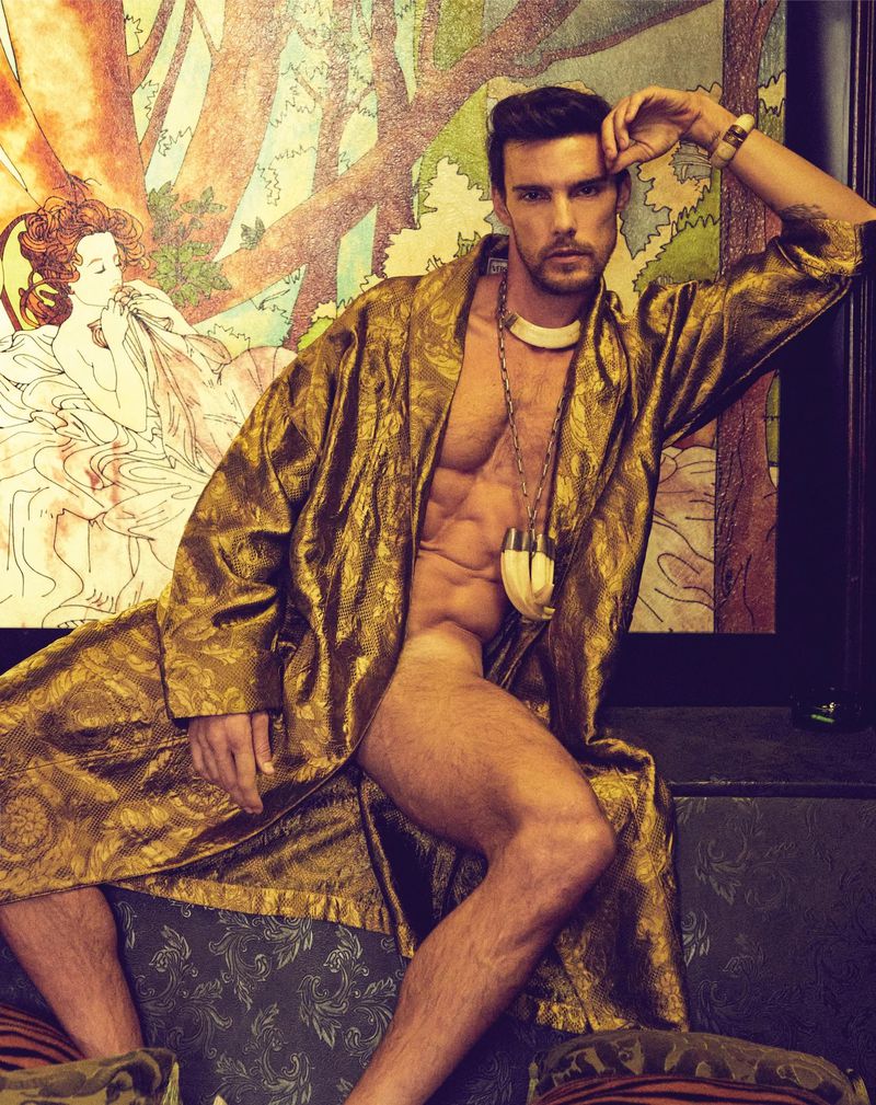 A muscular man wearing a gold robe is posed on a patterned couch in front of a drawing, showcasing a blend of art and physique.