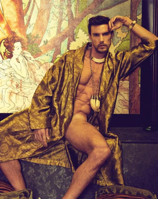 A muscular man wearing a gold robe is posed on a patterned couch in front of a drawing, showcasing a blend of art and physique.