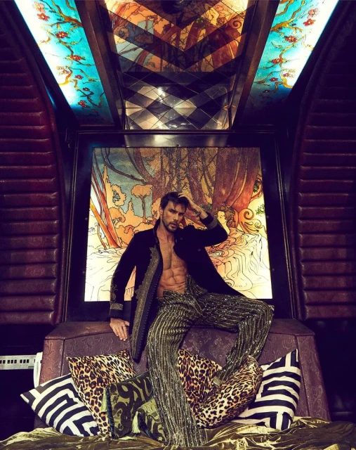 A male fashion model poses on a bed with decorative pillows wearing an unbuttoned black jacket and gold-striped pants in an indoor setting.