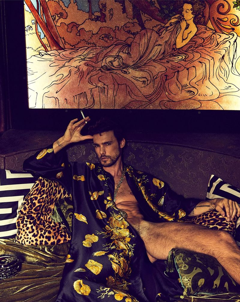 An adult male with dark hair, wearing a floral robe, poses on a couch with decorative pillows beneath an art nouveau style painting.