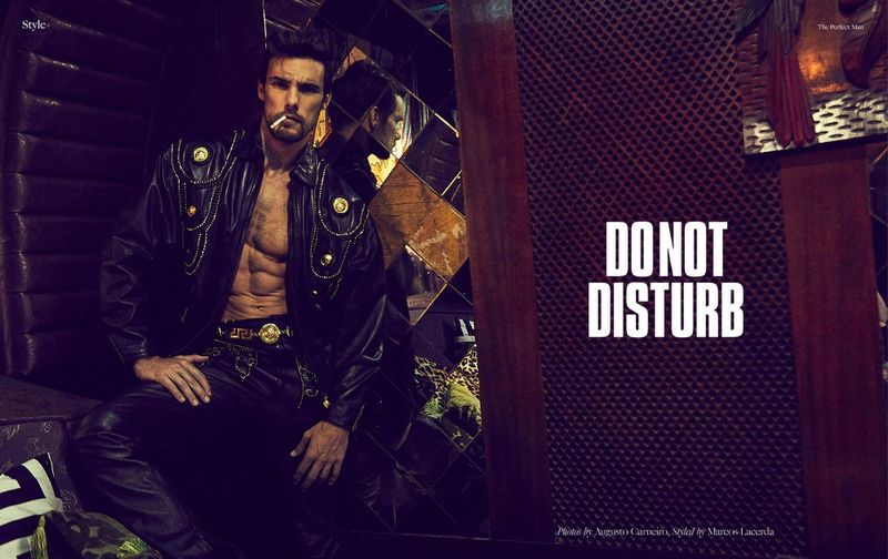Stylish adult male fashion model in leather outfit posing indoors with a cigarette, wearing jewelry, in a dark interior.