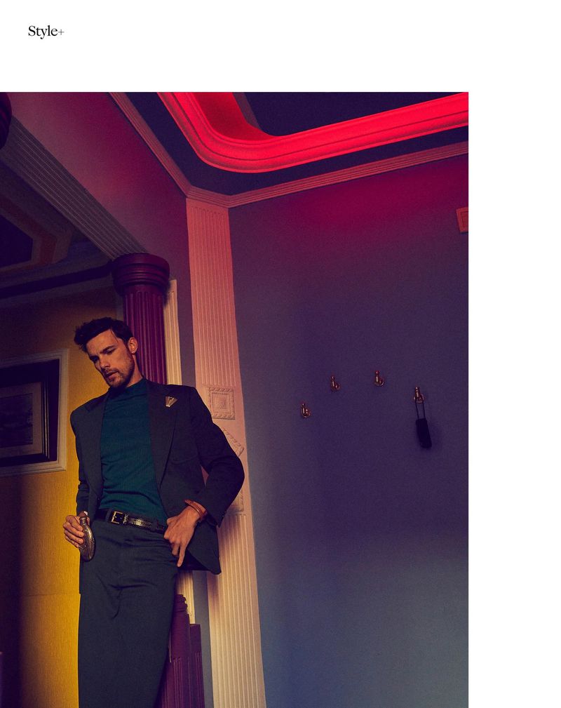 A man in a suit poses against a wall with a red neon light, wearing a dark green suit and holding a flask, in a dimly lit indoor setting.