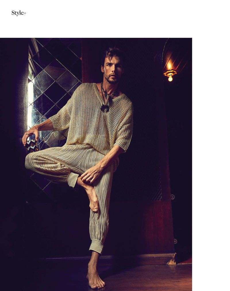 A man in a neutral-tone knitted top and pleated trousers stands on one leg in a dimly lit interior setting with a reflective wall.