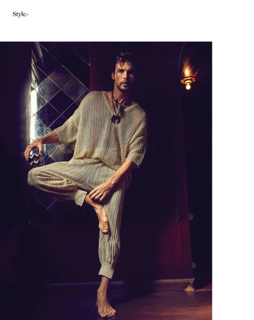 A man in a neutral-tone knitted top and pleated trousers stands on one leg in a dimly lit interior setting with a reflective wall.