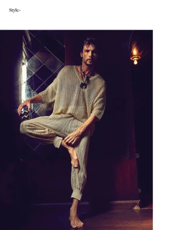 A man in a neutral-tone knitted top and pleated trousers stands on one leg in a dimly lit interior setting with a reflective wall.