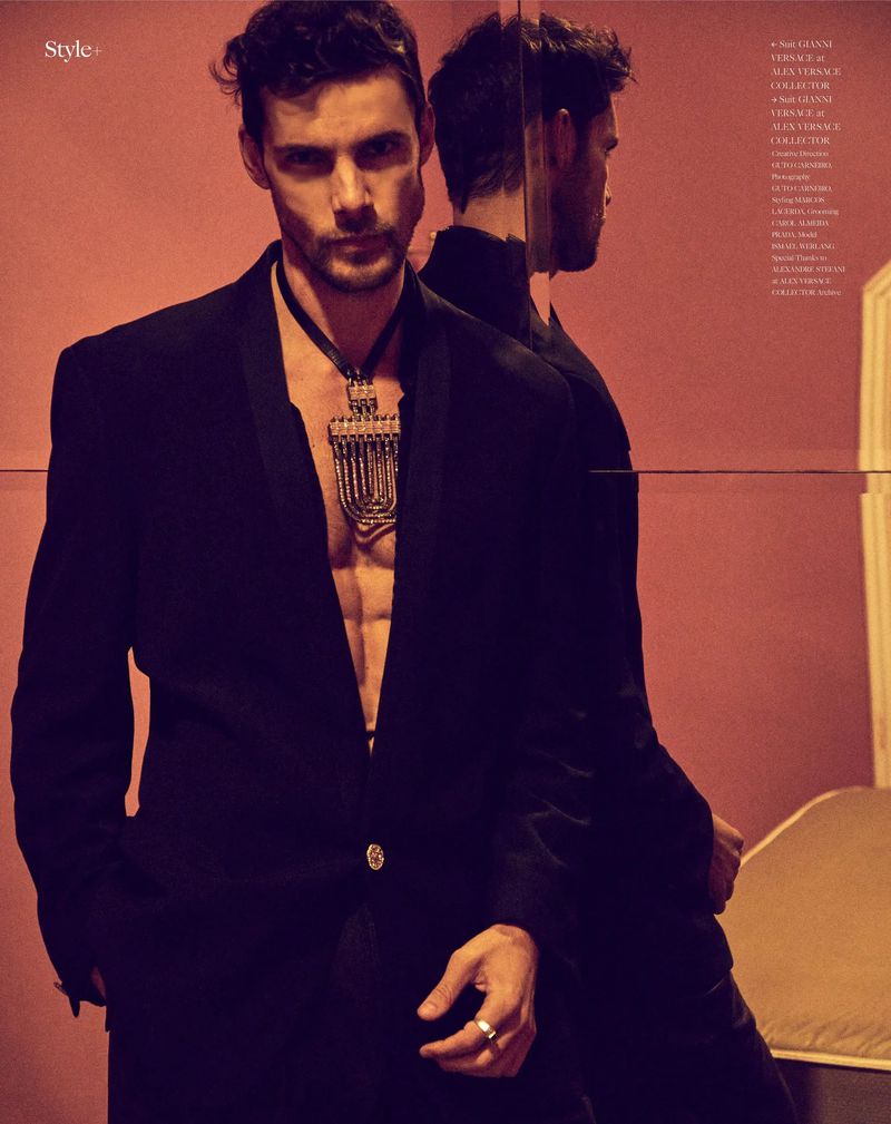 Male fashion model poses indoors wearing a black blazer and statement necklace, reflected in mirror against a salmon-colored wall.