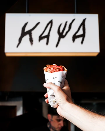 Hand holding a gyro wrapped in branded paper with visible tomato slices, against a dark background and a lit sign with black text.