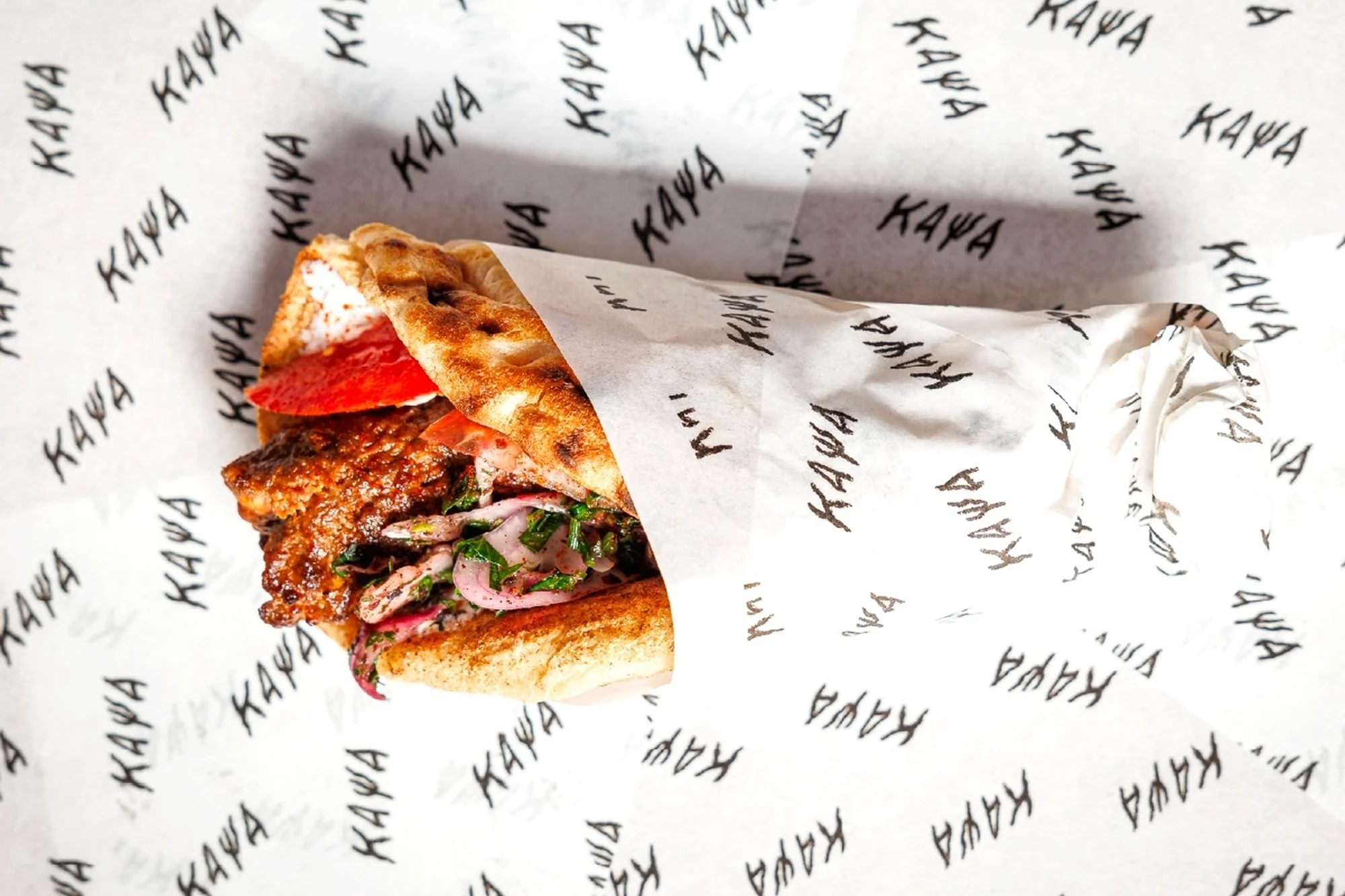 Close-up shot of a sandwich filled with meat, tomato, and onions, wrapped in logo-printed paper, placed on a matching logo background.