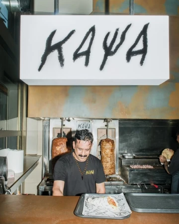 Man with mustache stands behind a counter with food on a tray in front of a restaurant grill with meat rotating. 