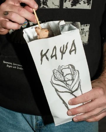 Adult holding a paper bag with a charcoal drawing on it and a snack on a stick emerging from the bag, on a dark background.