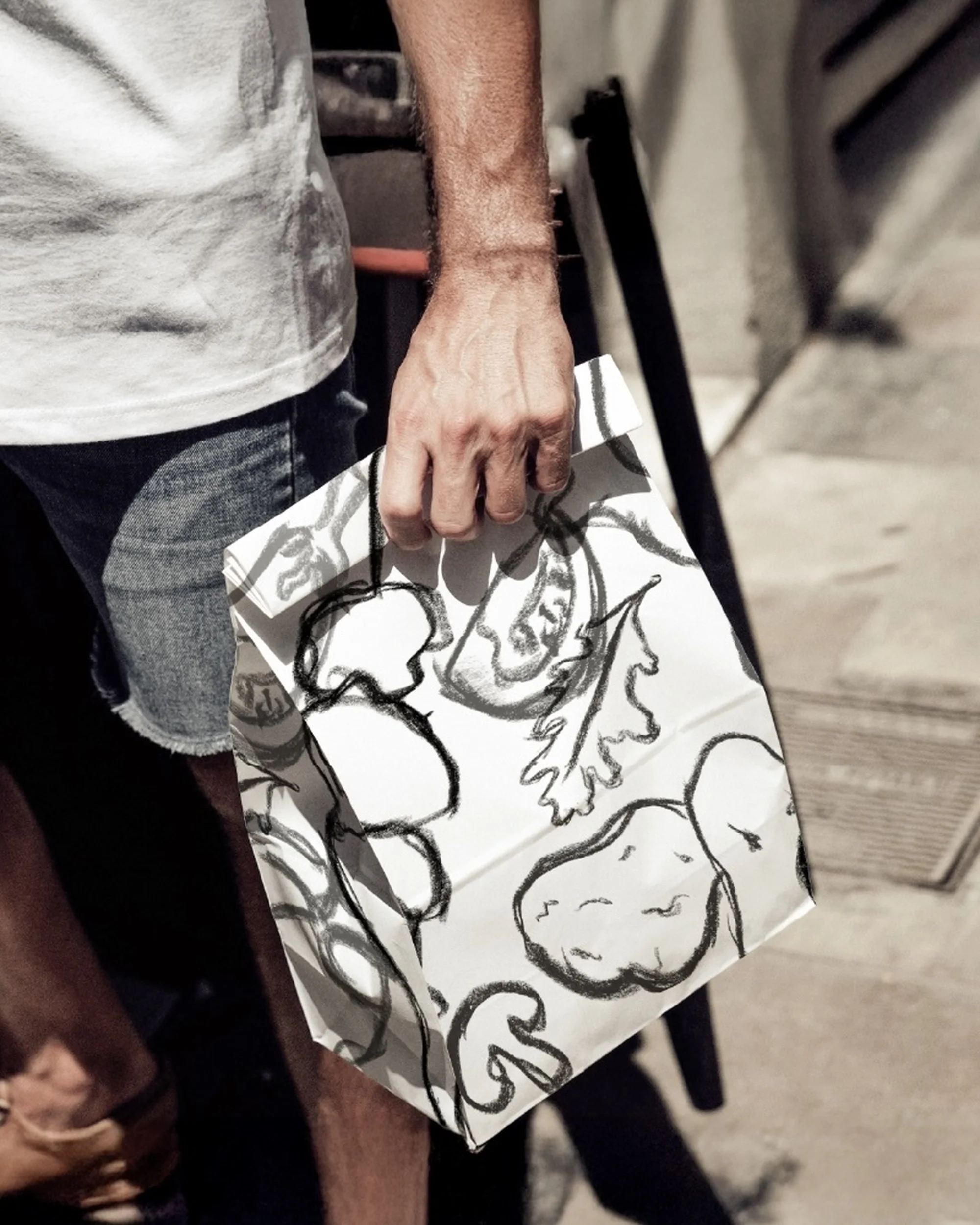Man carries a white paper bag with sketched vegetables. The bag features a drawing of a potato, mushroom, tomato, and lettuce.