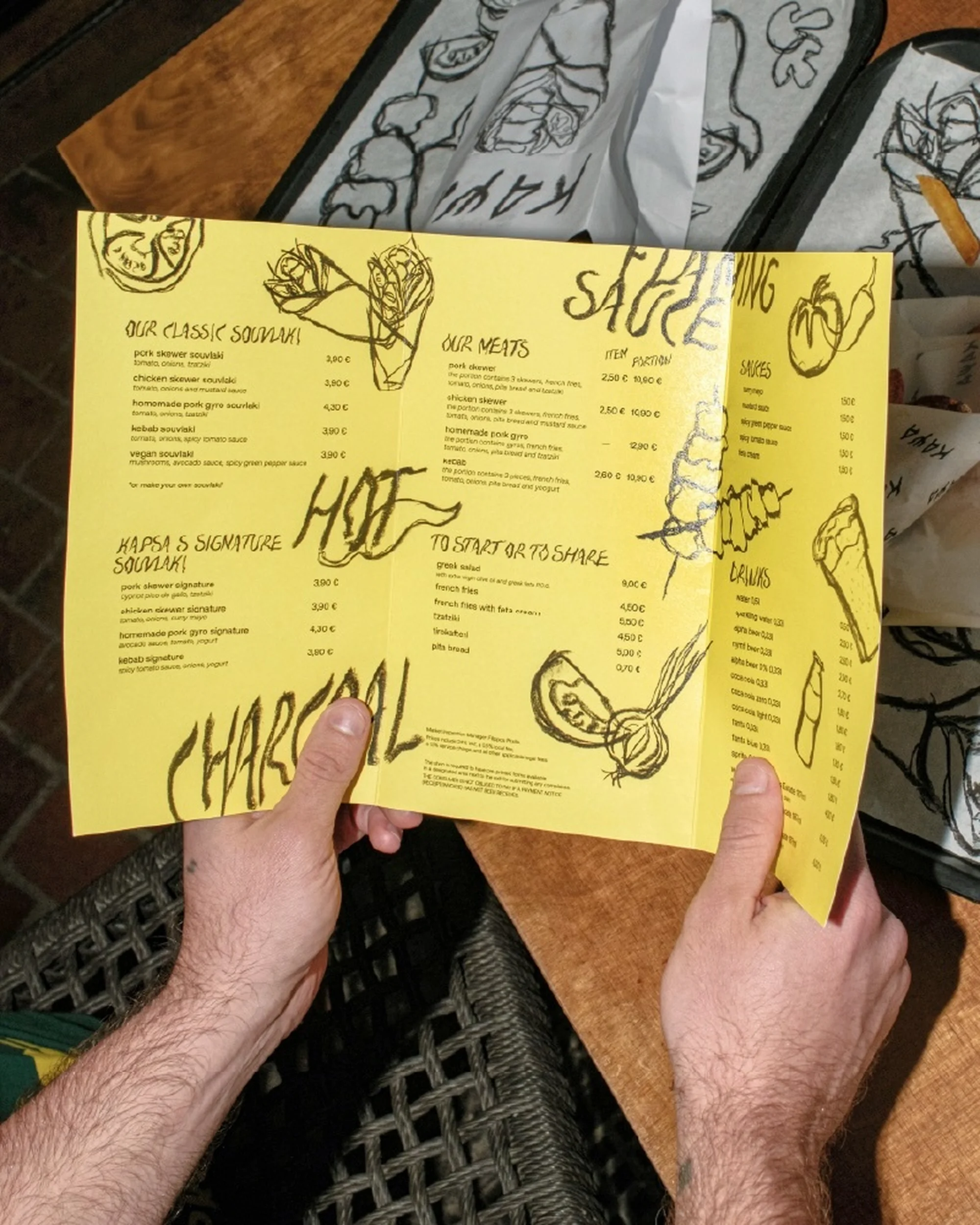 Man holds yellow menu with black lettering and illustrations. The menu lists a variety of foods and beverages. Table setting.