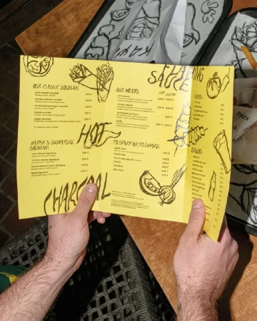Man holds yellow menu with black lettering and illustrations. The menu lists a variety of foods and beverages. Table setting.