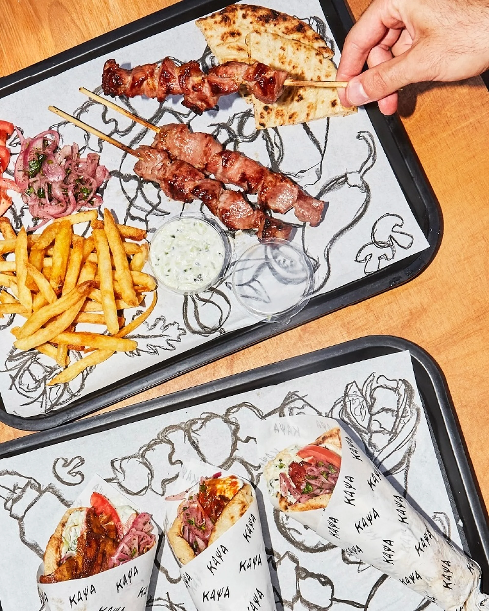 Two trays present an array of Greek food: meat skewers, gyro wraps, fries, and tzatziki sauce, all served indoors on paper liners.