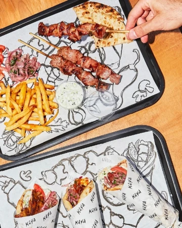 Two trays present an array of Greek food: meat skewers, gyro wraps, fries, and tzatziki sauce, all served indoors on paper liners.
