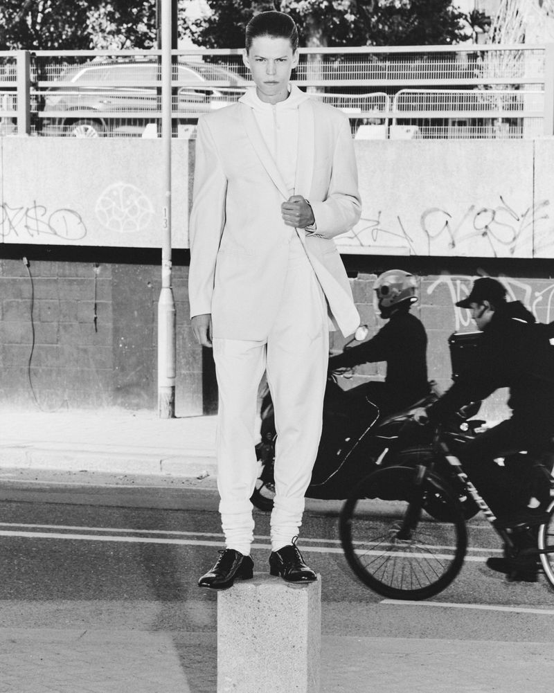 Monochrome fashion shot of a young adult man posing on a block in a city street with traffic and a graffitied wall in the background.