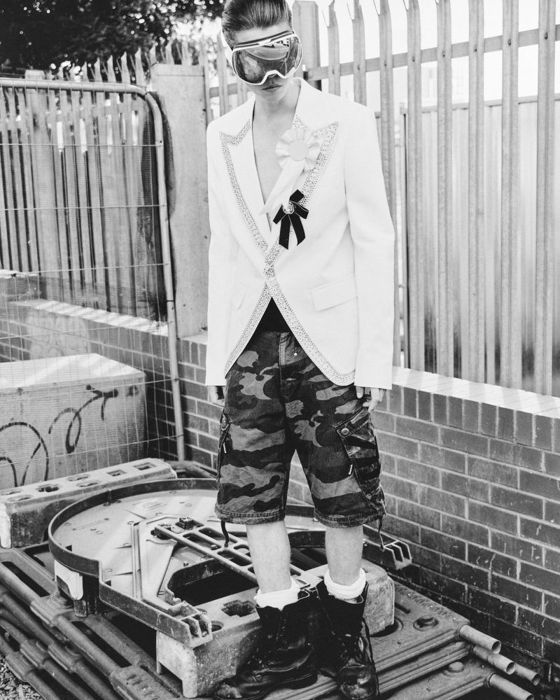 A male fashion model poses outdoors wearing futuristic ski goggles, camouflage shorts, white socks, and black boots, next to scaffolding.