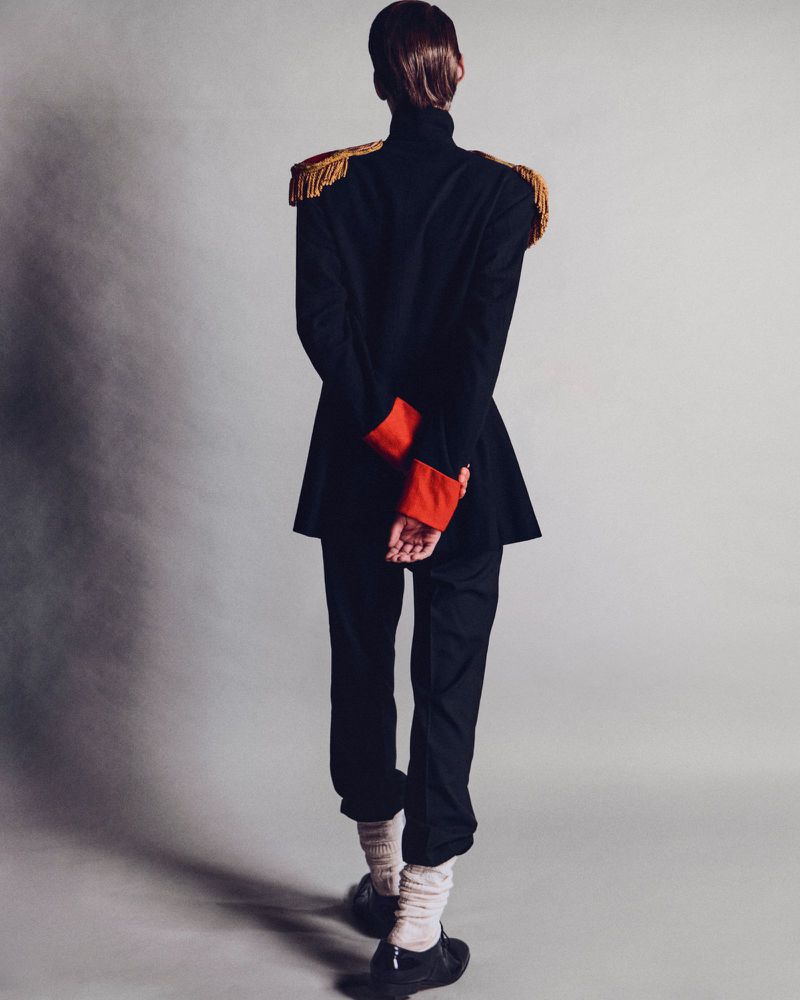 Back view of a person in a dark jacket with gold fringed epaulettes, black pants, and shoes standing on a gray studio backdrop.