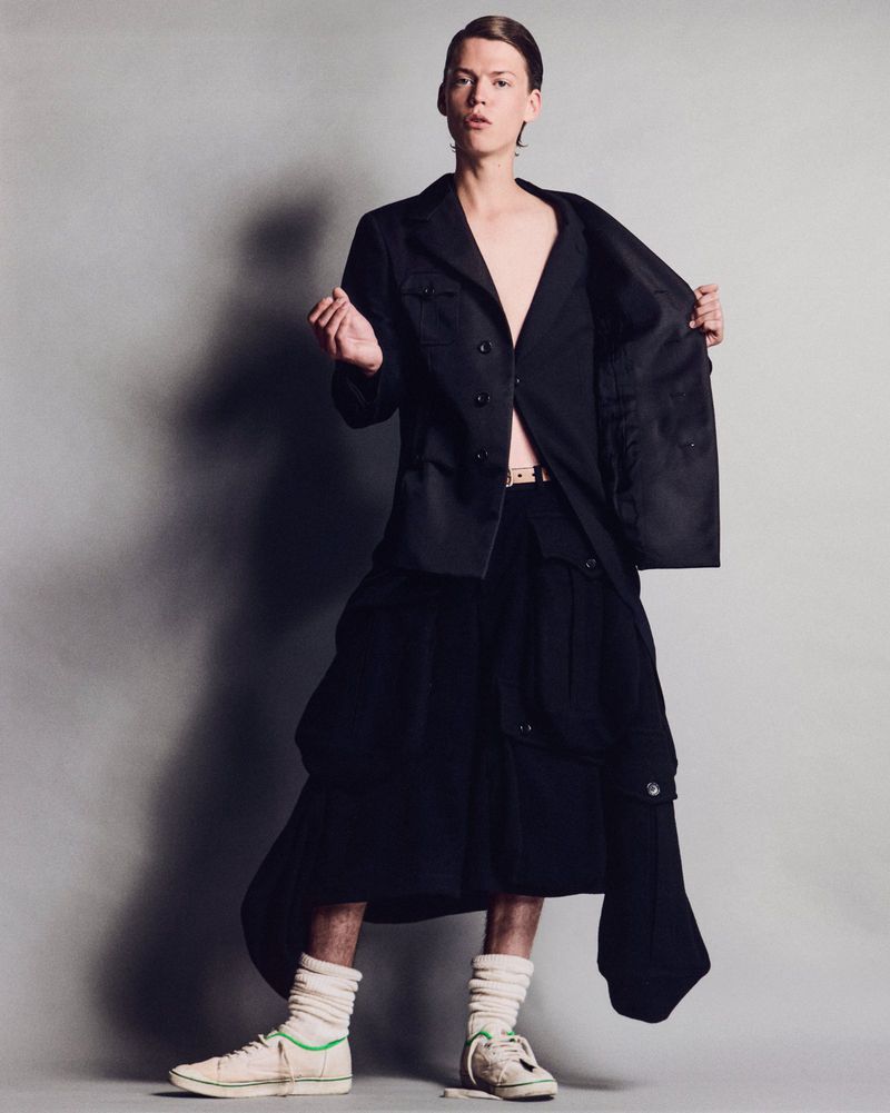 Fashionable male in a black jacket and skirt poses indoors against a grey background, showcasing a modern editorial look.