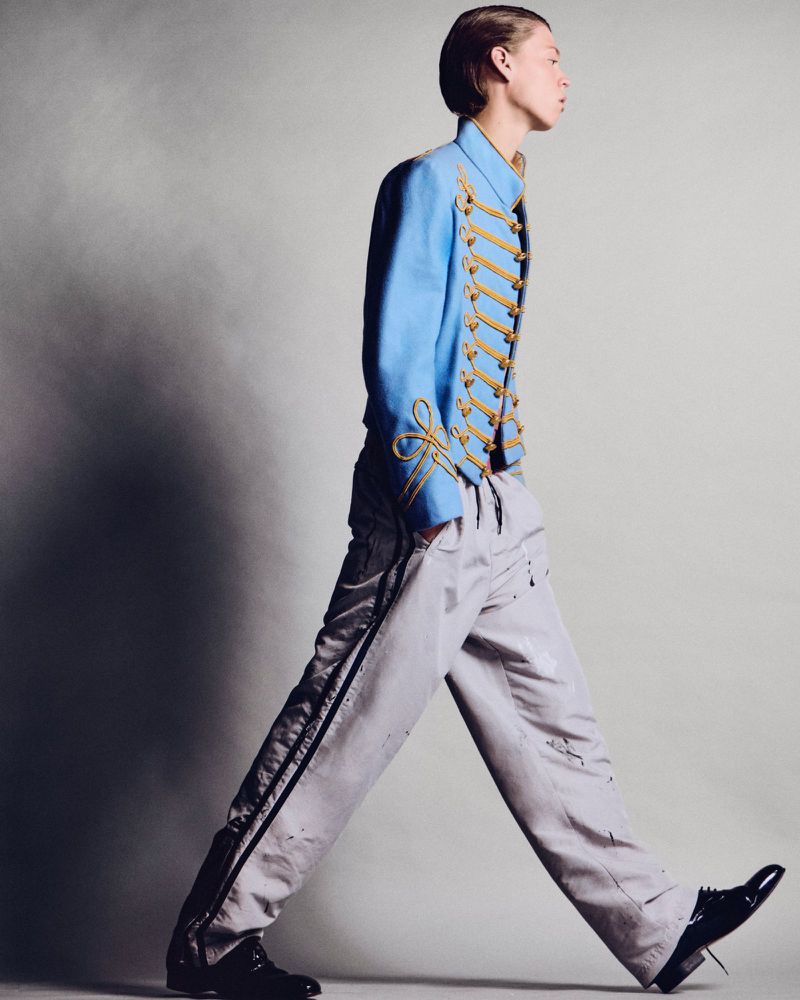 A person in a blue military style jacket and grey pants walking on a grey background in a fashion style portrait shot.