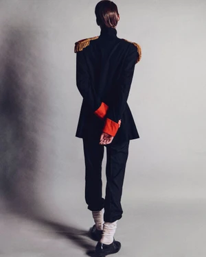 Model standing with back to camera wearing a black military style jacket with gold fringe epaulettes and red cuffs