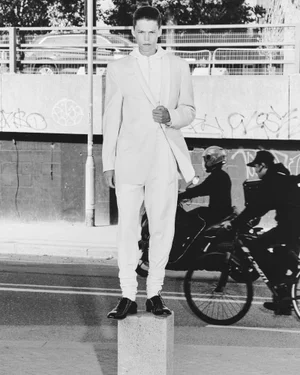 Monochrome fashion editorial featuring a standing male model balancing on a stone block against an urban street background with passing traffic.