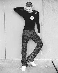 Young man in dark long-sleeve top and textured trousers posing against a plain wall for Parachute Magazine editorial Ruben.