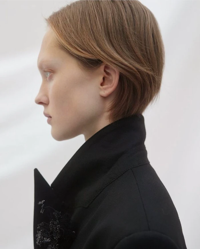 Profile portrait of a model with short brown hair wearing a black jacket against a plain white background.