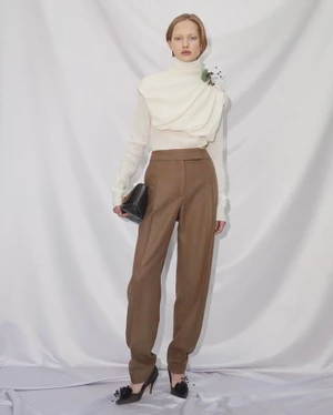 Model in cream turtleneck and brown trousers holding a black handbag against a white seamless backdrop