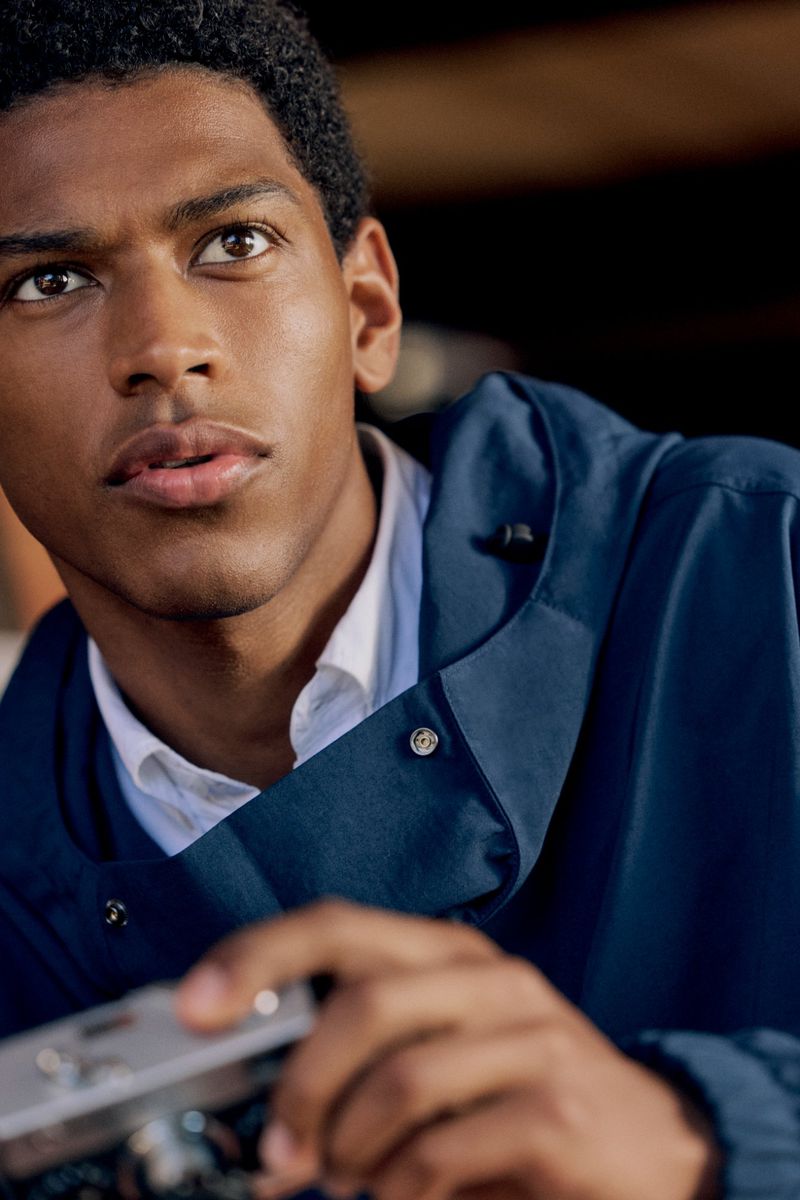 Portrait of an African-American male wearing a blue jacket and holding a camera, looking off-camera to the upper left of the frame.