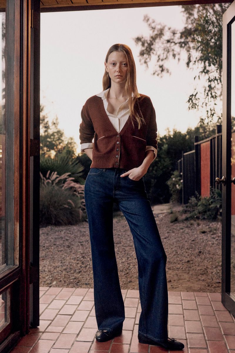 A slender, fair-skinned adult woman stands in a doorway wearing a brown cardigan, blue jeans, and black shoes in an outdoor setting.