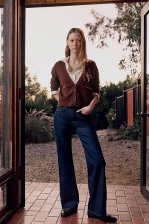A slender, fair-skinned adult woman stands in a doorway wearing a brown cardigan, blue jeans, and black shoes in an outdoor setting.