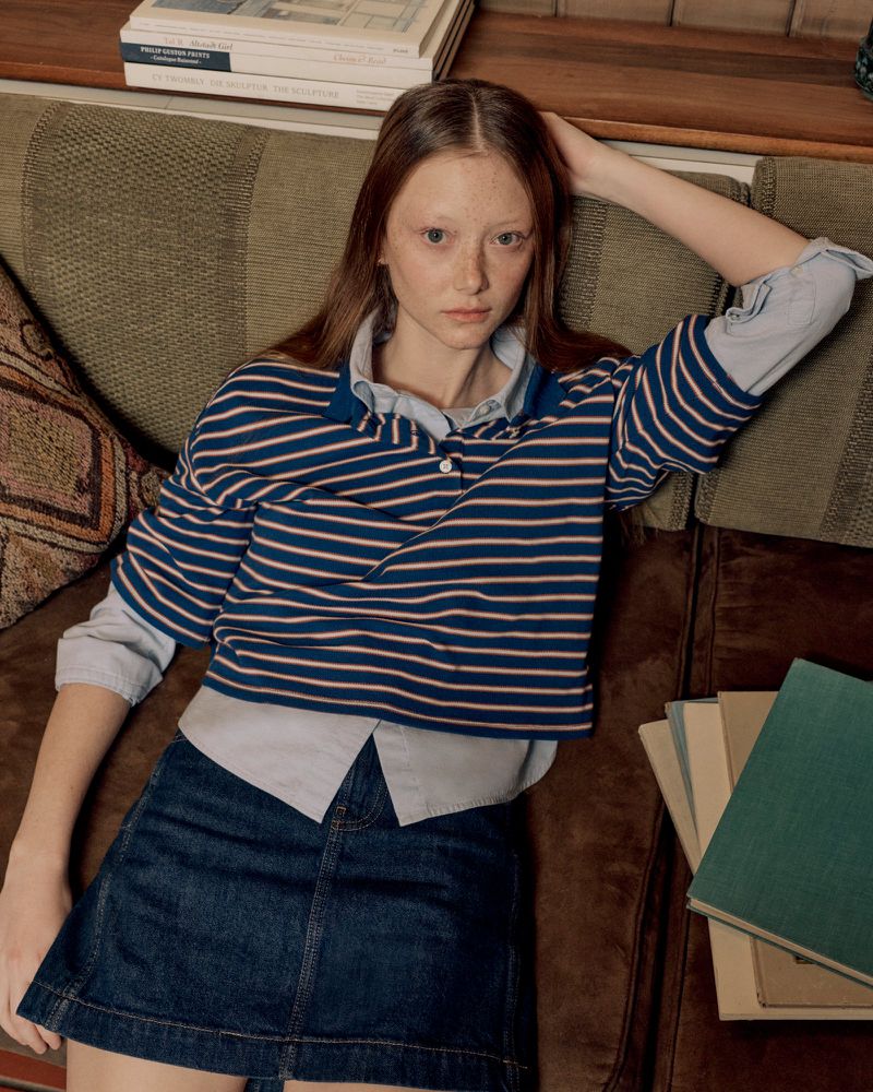 Young woman poses on a brown striped couch wearing a blue and white striped collared shirt and a denim skirt in an indoor setting.