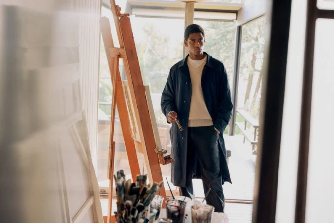Young man in casual wear stands near easel inside a modern home, paintbrush in hand, illuminated by natural light.