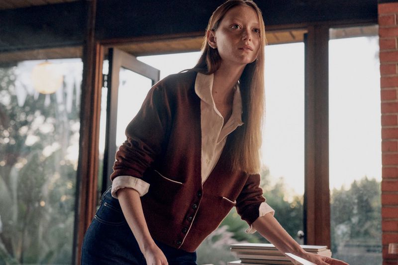 Woman in cardigan standing indoors near natural light, wearing jeans and a white shirt, arranging a stack of paper and books.