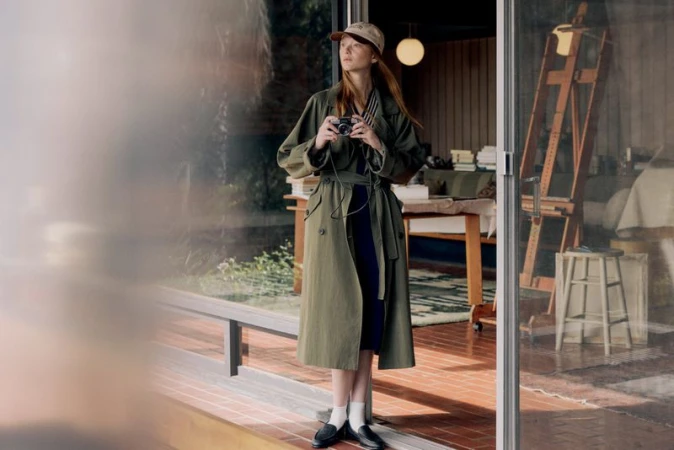 A stylish young woman stands near a glass door, holding a camera and wearing an olive trench coat, a cap, and black loafers with white socks.
