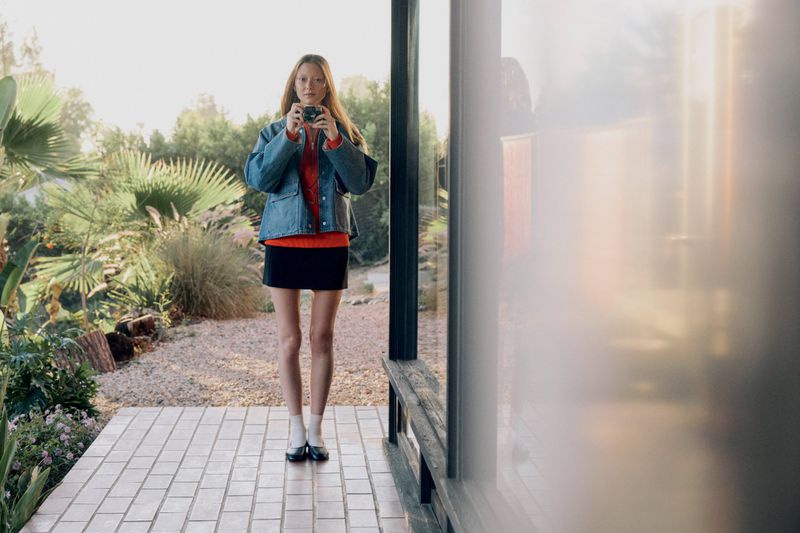 Adult female taking photo with a camera outside a building with greenery in the background, wearing a denim jacket and skirt.
