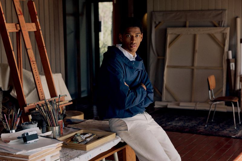 Young adult male in blue jacket poses in art studio with painting easel, brushes, canvases, and painting supplies on table in daytime.