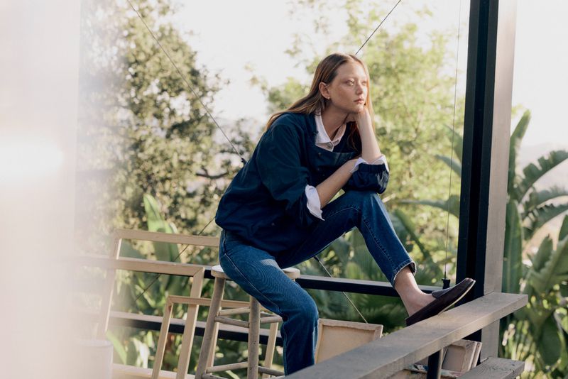 A woman sits on a ledge overlooking a yard, wearing a dark blue jacket and blue jeans, with trees visible in the background on a sunny day.