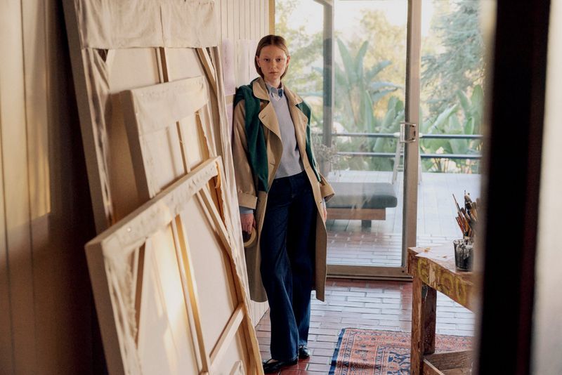Woman in trench coat poses in light-filled indoor space near stacked canvas frames, with lush green trees visible through a glass door.