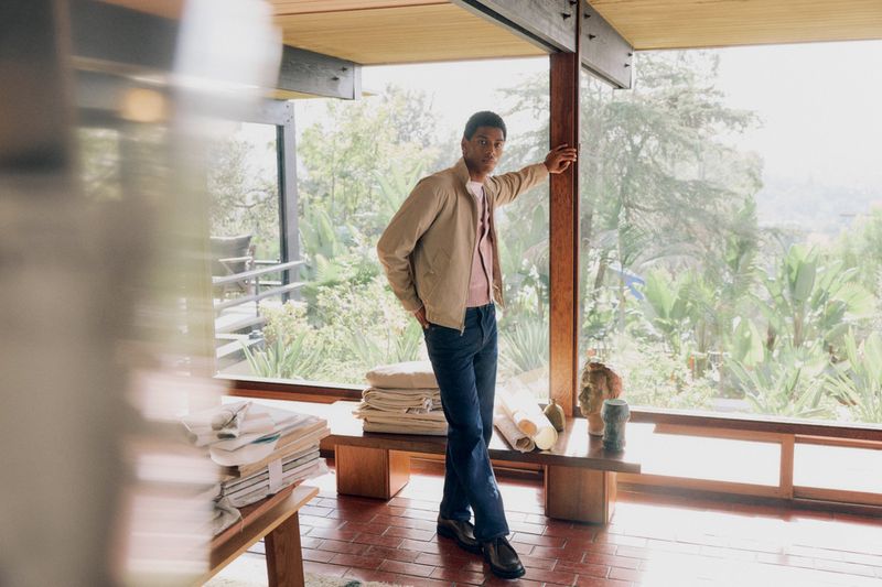 A man in a jacket stands in a modern room with large windows, gazing out at a leafy landscape, with a table and objects in the foreground.