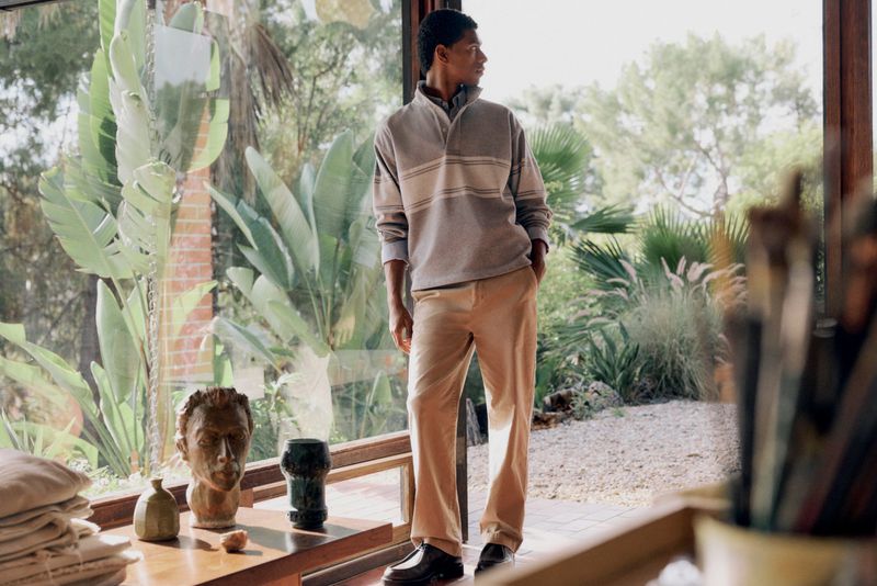 A dark-skinned man stands indoors near a window wearing a grey, cream, and brown striped collared shirt and tan trousers in a fashion editorial.